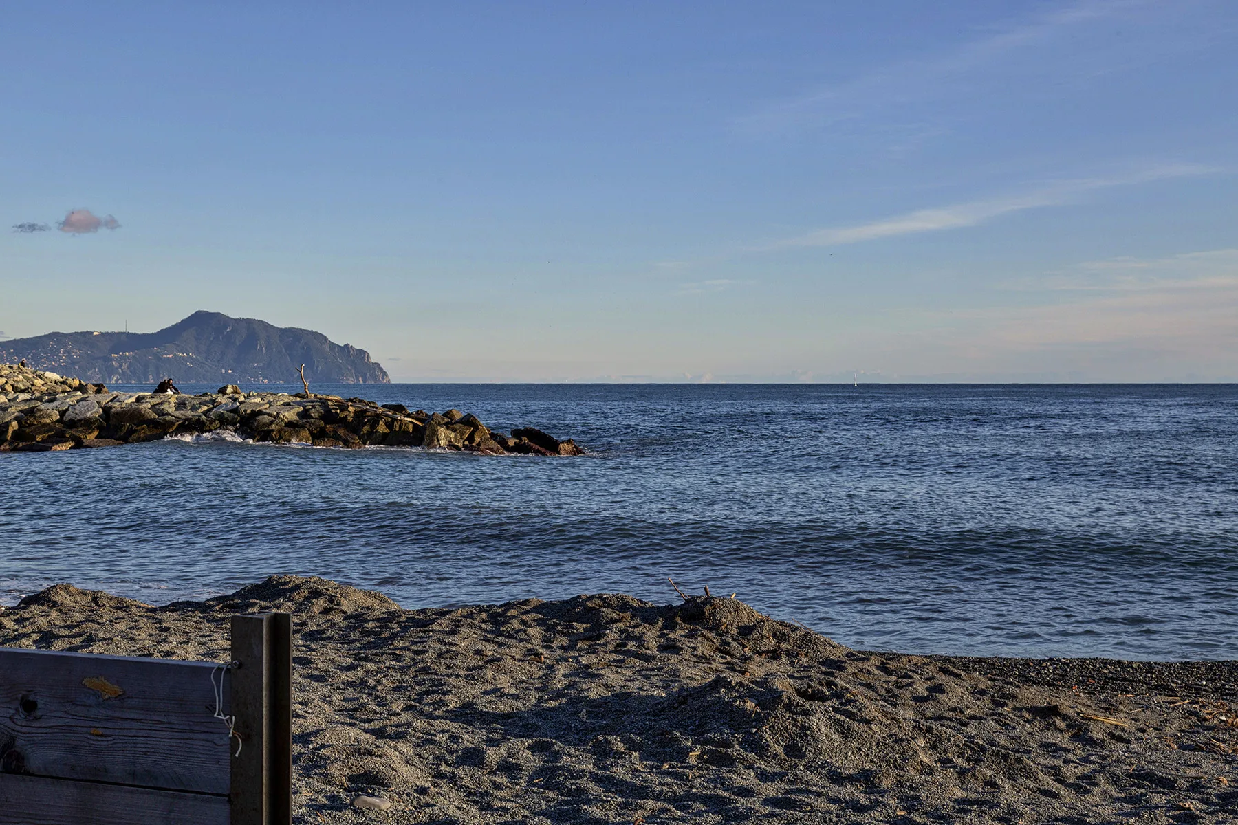 A rocky shoreline meets calm water, with mountains visible along the coast.