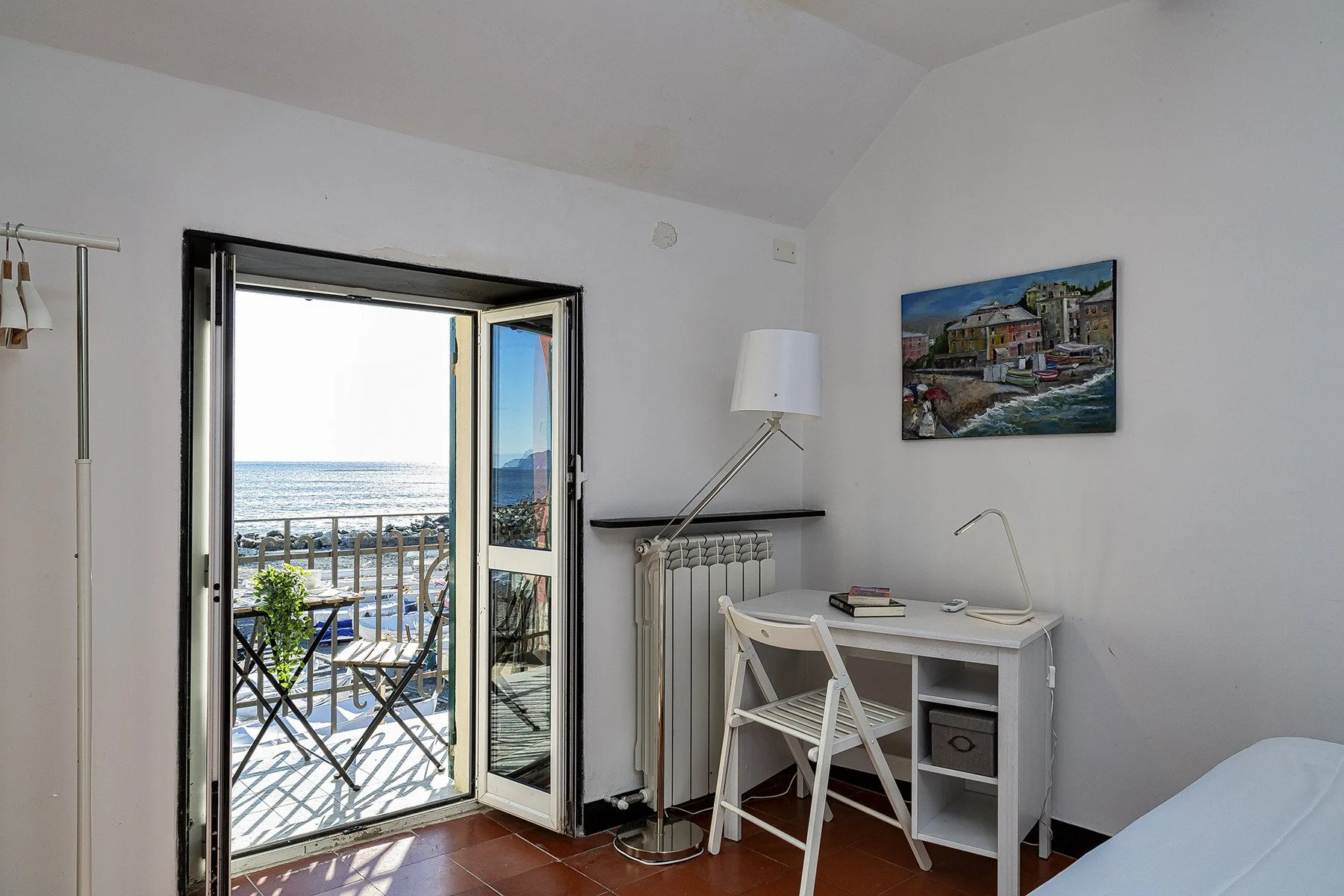 A bright room opens onto a balcony overlooking the sea, with a desk beside the doorway.