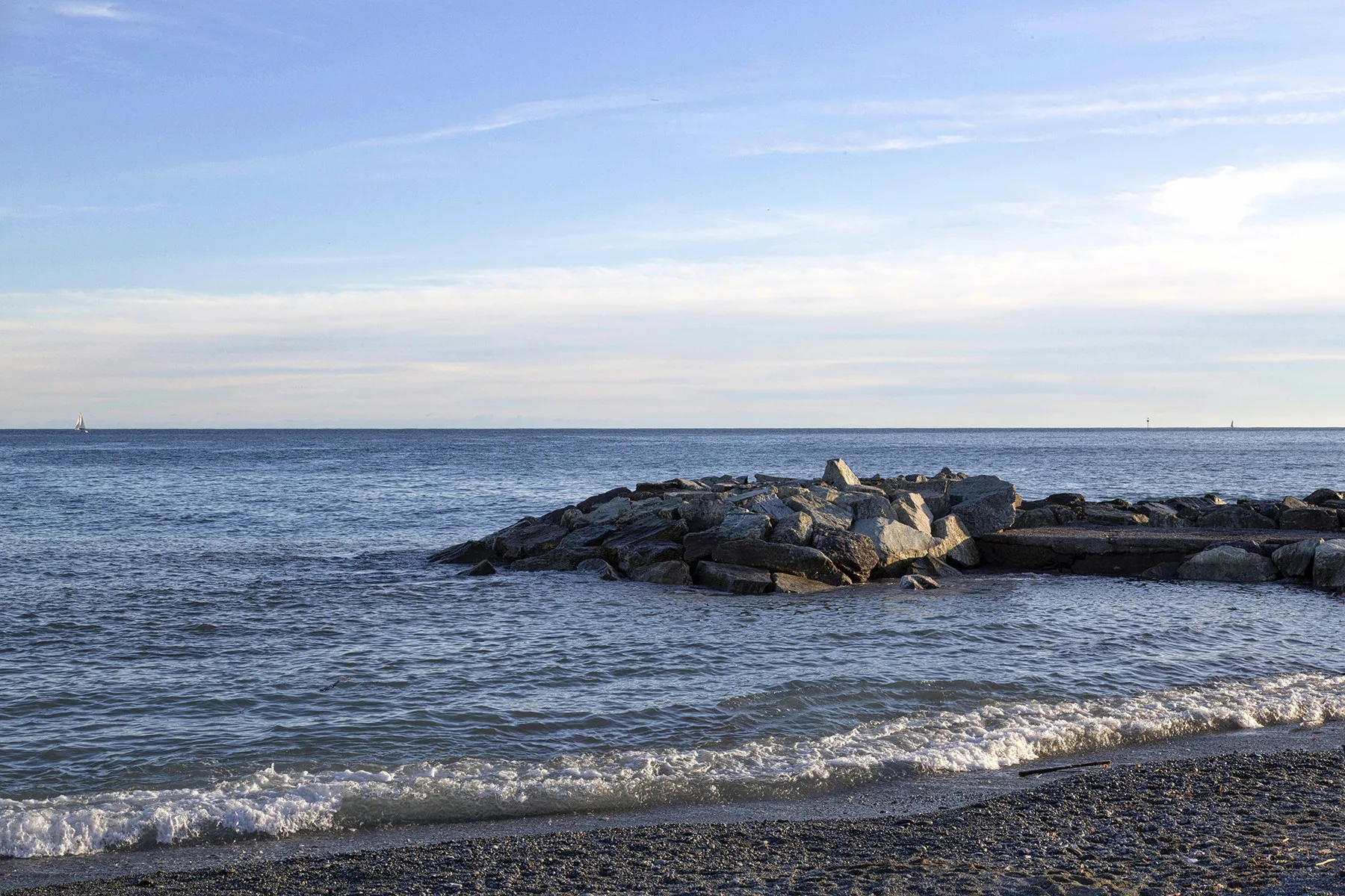 A pebbled beach meets calm water beside a rocky breakwater.