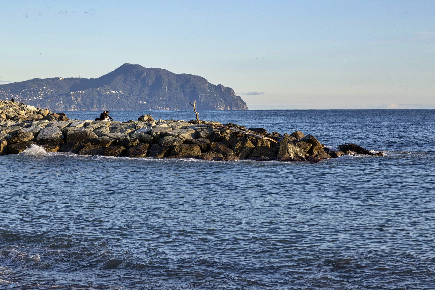 A rocky breakwater extends into calm blue water, with mountains on the horizon.