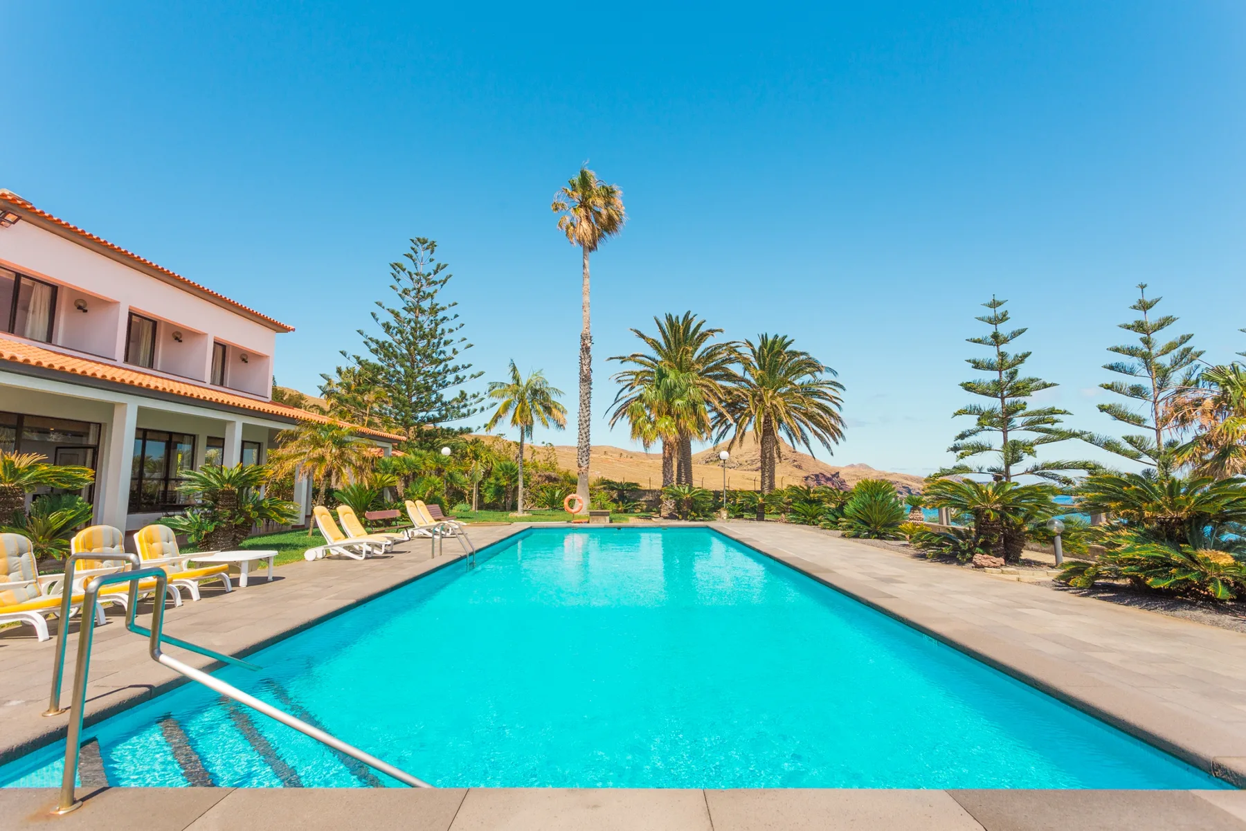 Una piscina junto a una casa grande, rodeada de palmeras y asientos al aire libre.