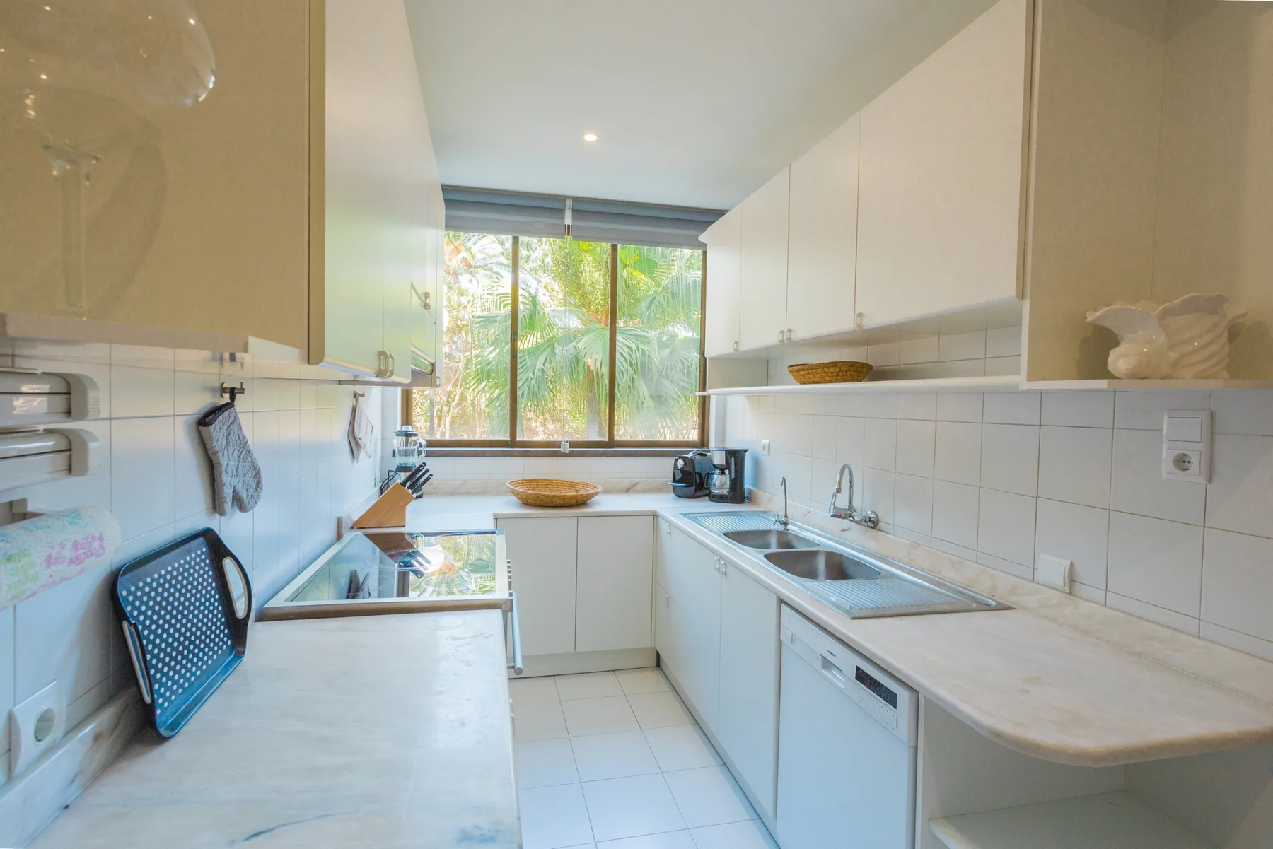 Una cocina luminosa con encimeras blancas y una ventana con vistas a la vegetación.