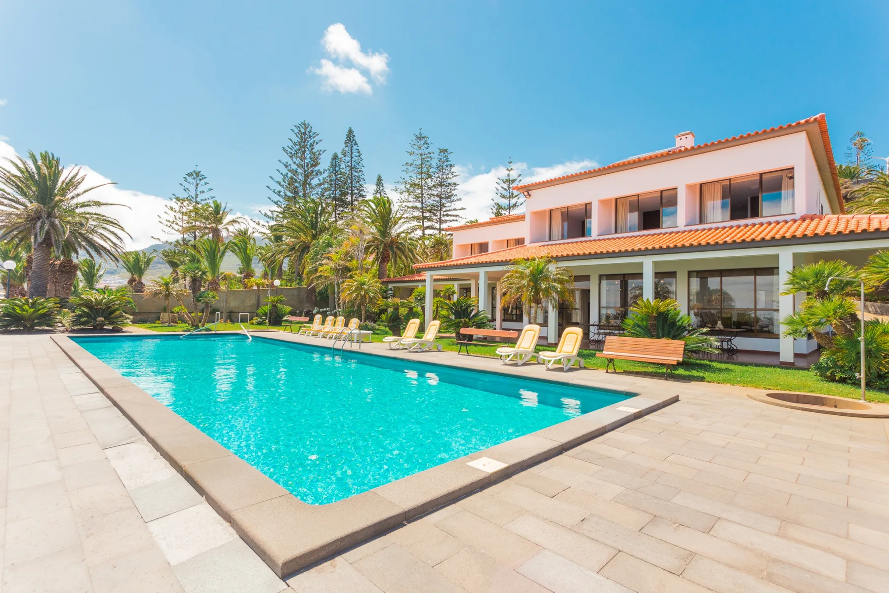 Una piscina junto a una casa grande, con palmeras y asientos en el patio.