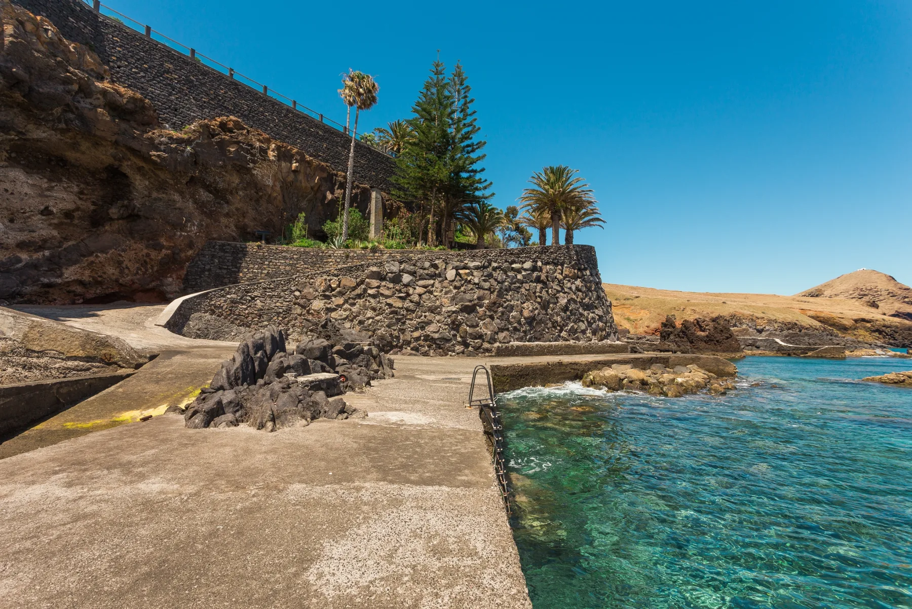 Una plataforma de hormigón junto al mar bordea aguas claras bajo una ladera rocosa con palmeras.