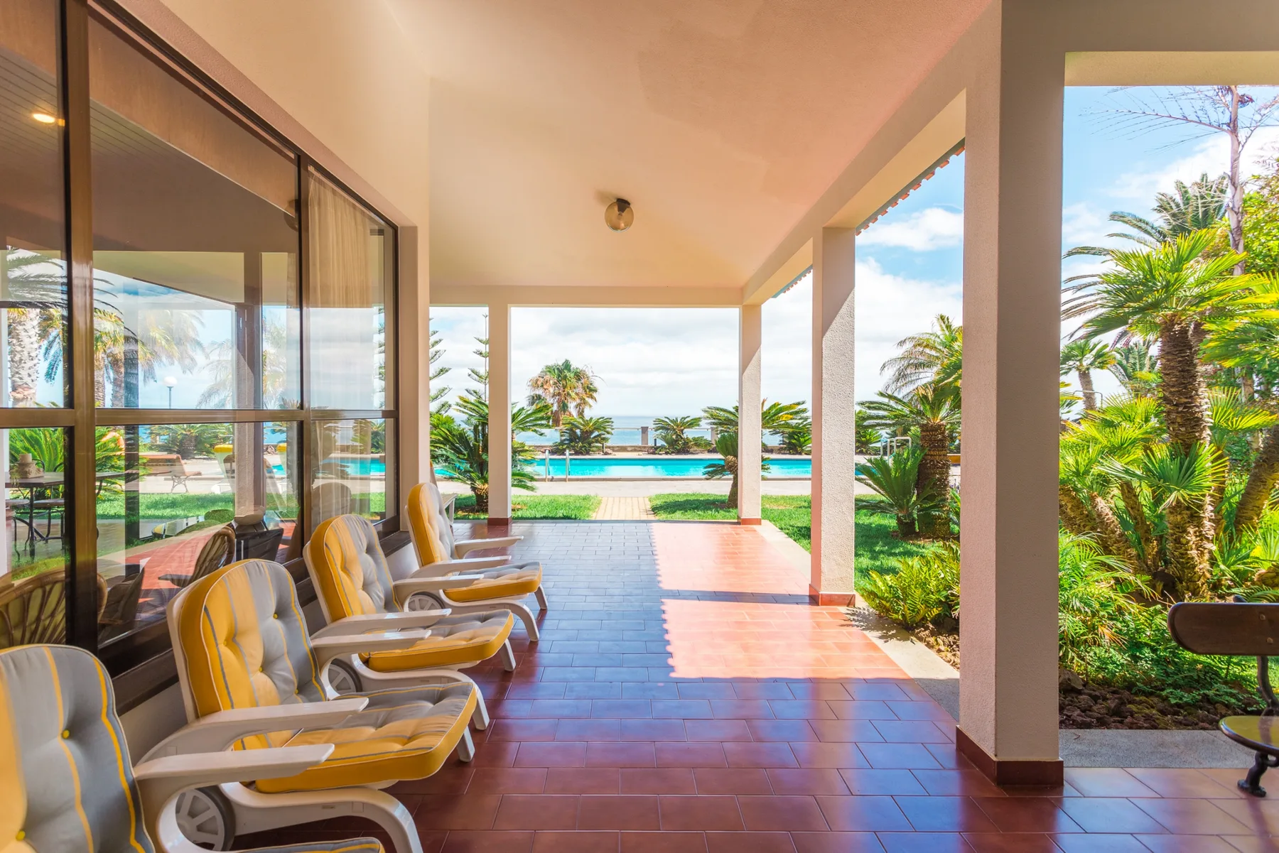 A covered patio with cushioned seating overlooks a swimming pool and tropical plants.