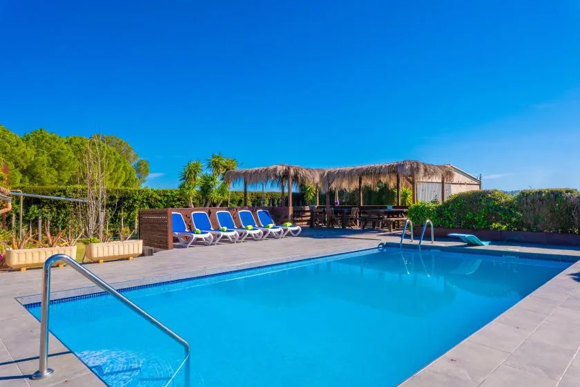 Una piscina junto a una terraza con asientos reclinables y un pabellón sombreado.