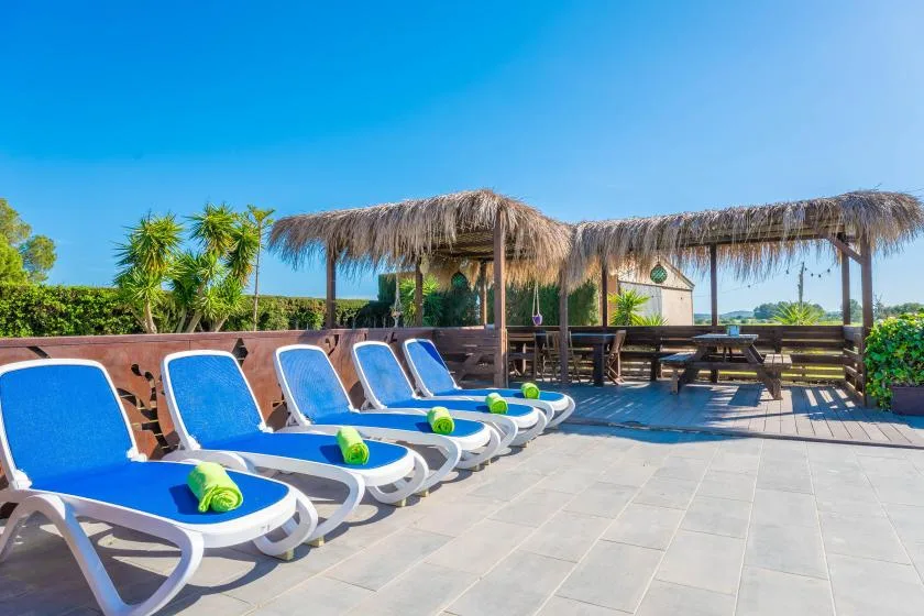Un patio soleado junto a la piscina, con tumbonas azules bajo un techo de paja.