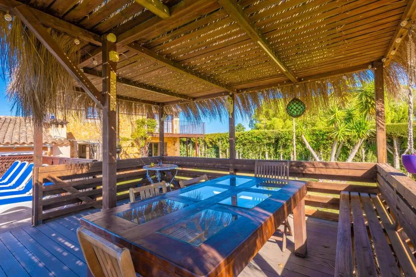Una zona de comedor exterior de madera está bajo una pérgola de paja junto a una piscina.