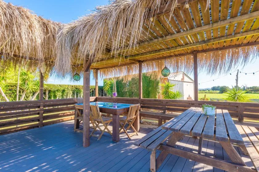 Una zona exterior de comedor de madera está bajo una pérgola de paja, con campo abierto al fondo.