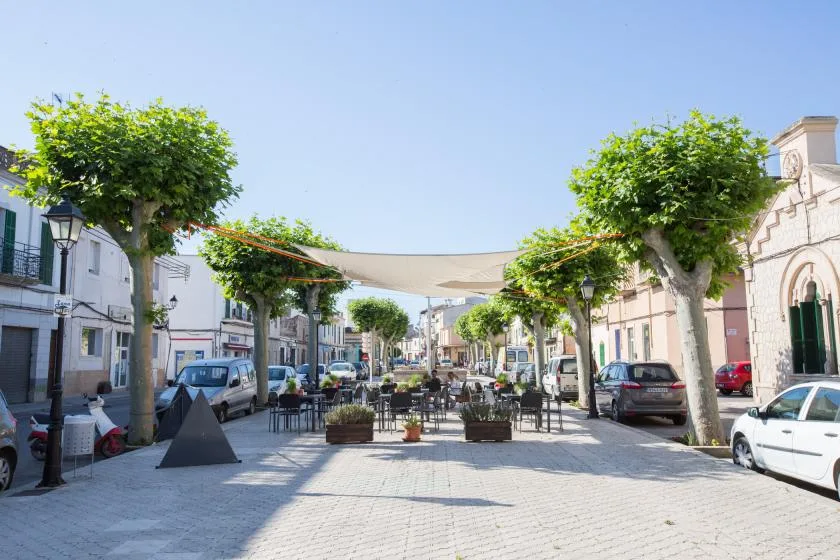 Una calle peatonal soleada con árboles frondosos, mesas al aire libre y un toldo.