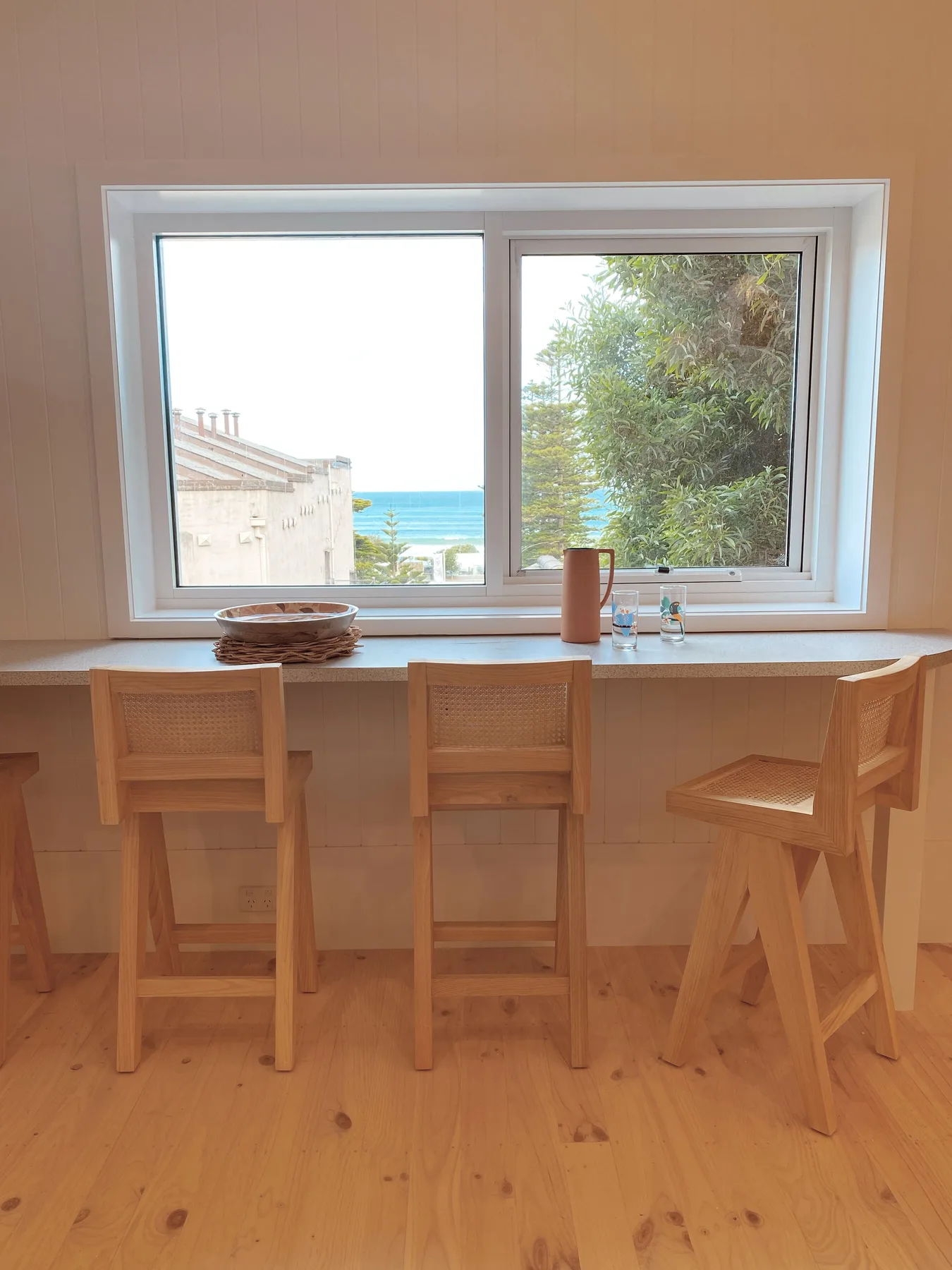 Una barra de madera con asientos da a una ventana con vista al mar.
