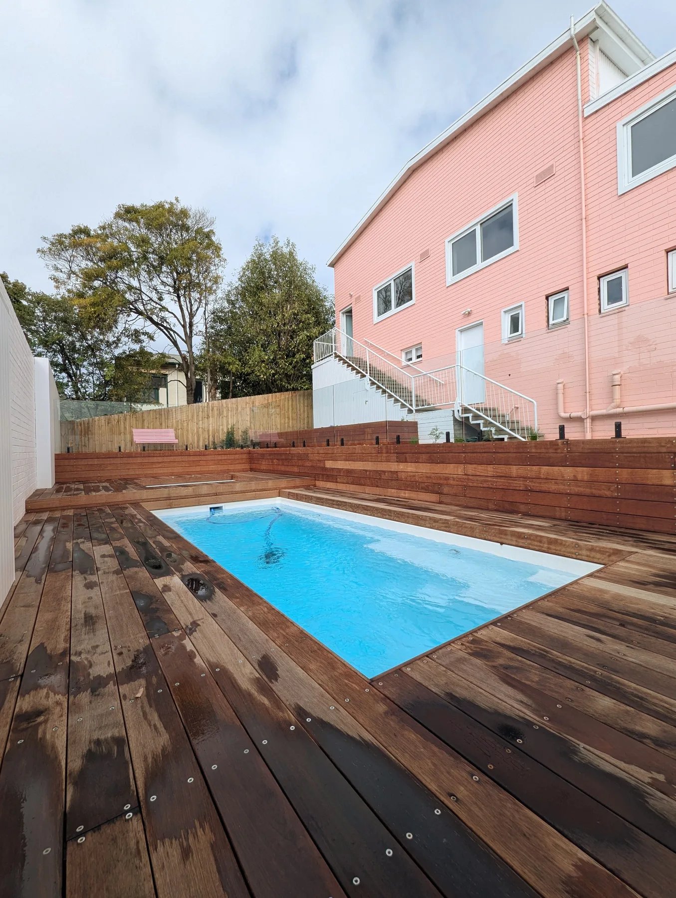 Una piscina al aire libre está rodeada por una terraza de madera junto a un edificio rosa.