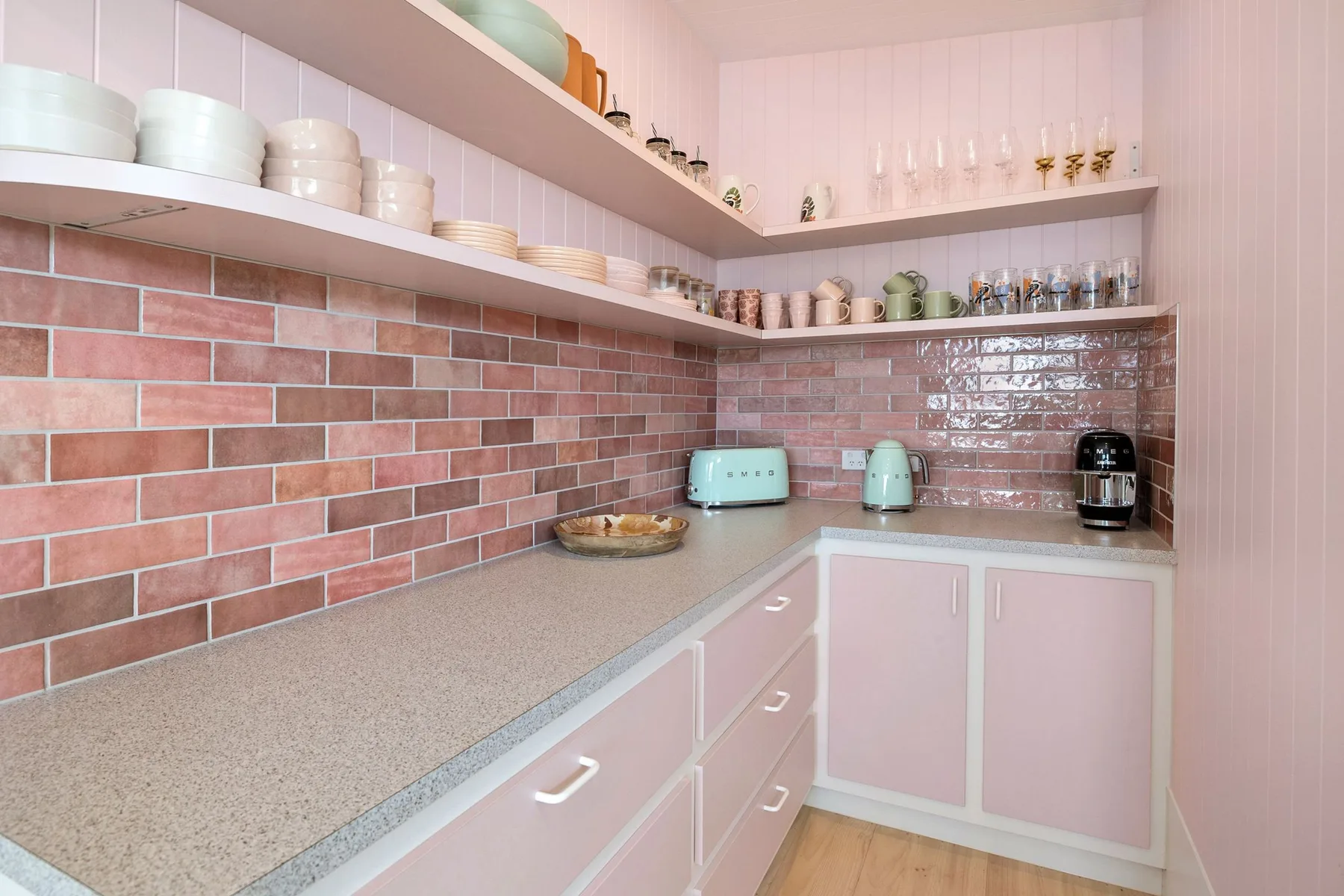 Una cocina rosa con estantes abiertos, pequeños electrodomésticos y un salpicadero de azulejos.