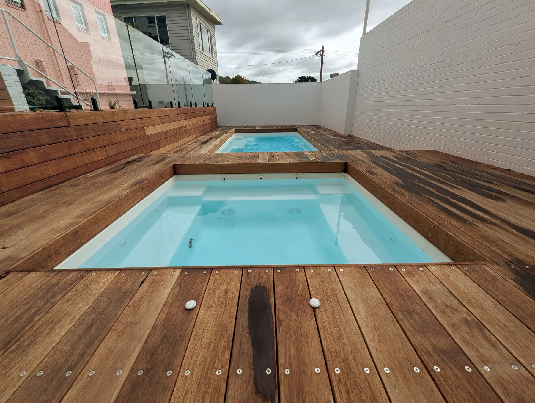 Dos pequeñas piscinas están integradas en una terraza de madera junto a una pared blanca.
