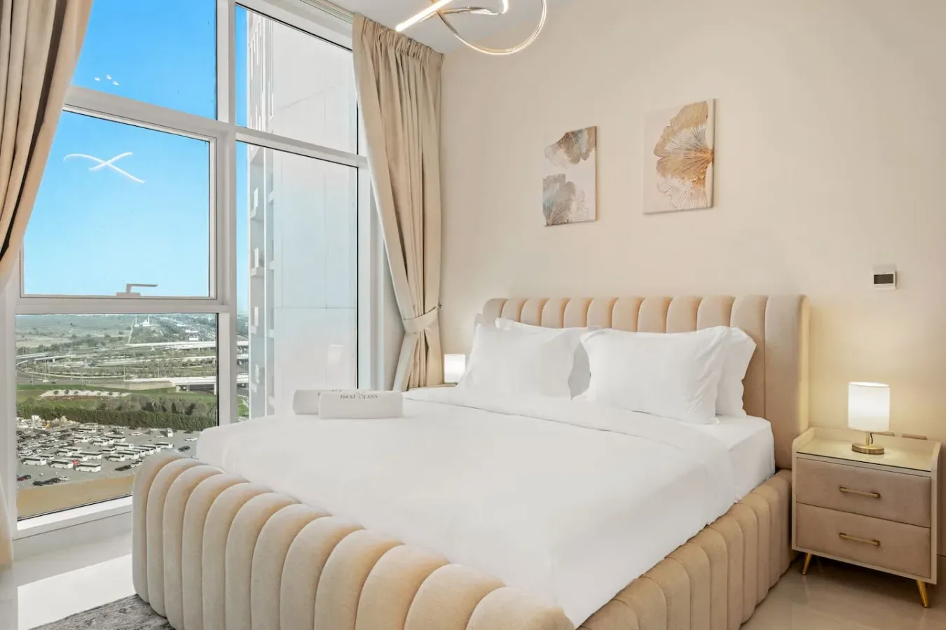 A bright bedroom with a white bed beside large windows and beige curtains.