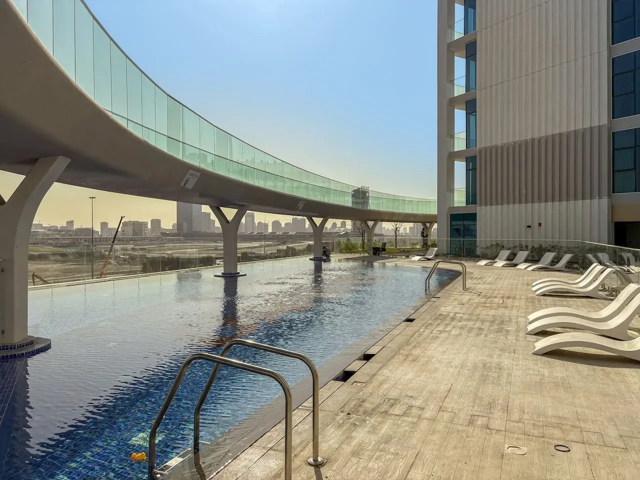 An outdoor swimming pool beside a sun deck with lounge seating and a curved glass walkway.