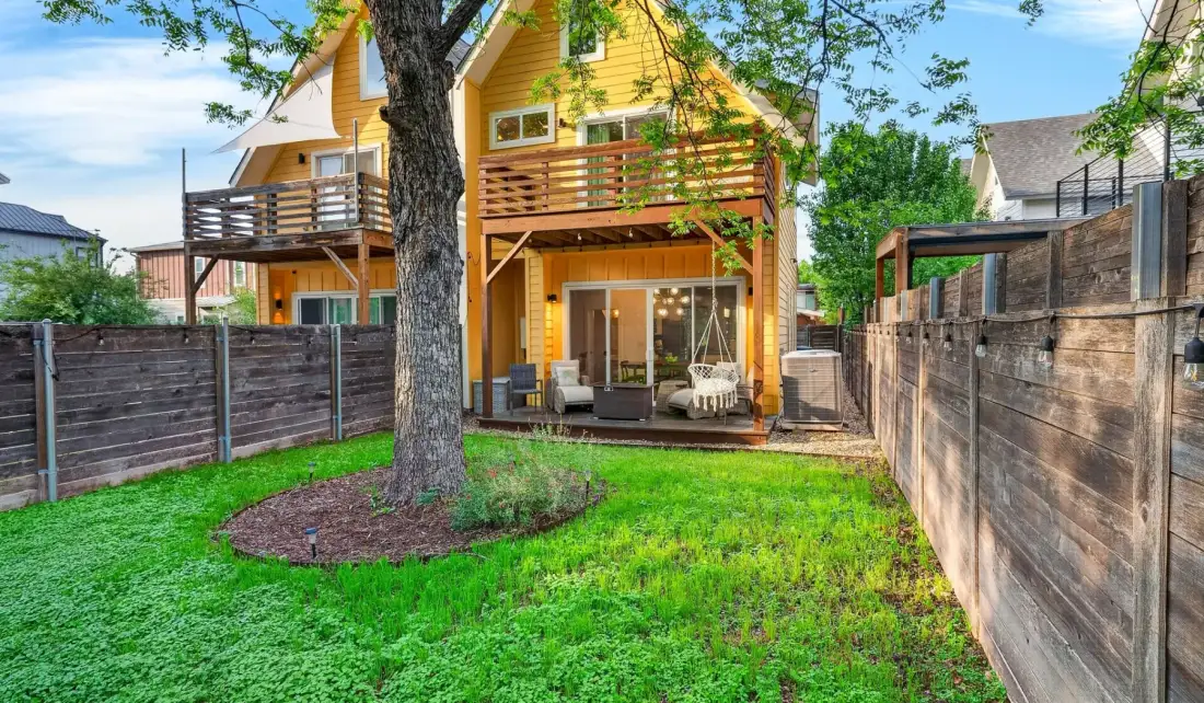 A yellow two-story house opens onto a fenced backyard with a deck and patio seating.