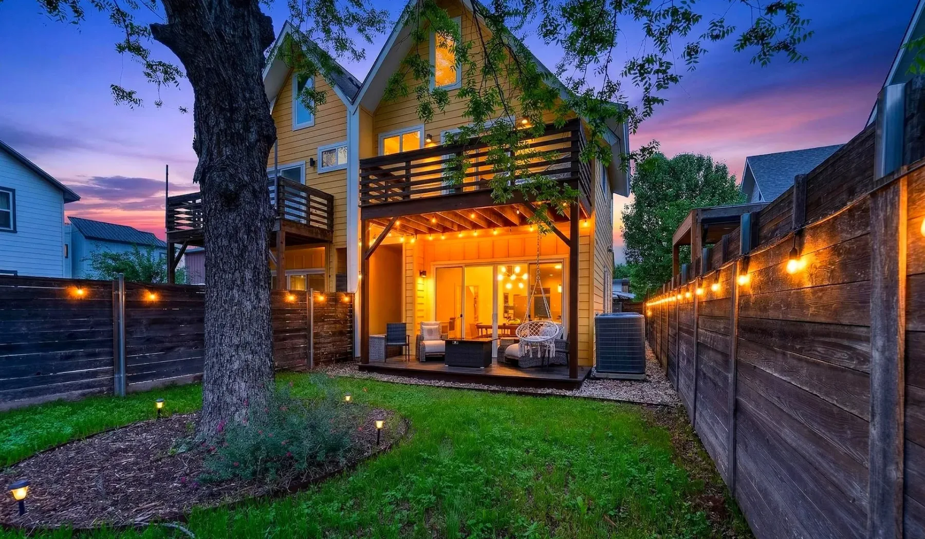 Una casa iluminada da a un patio cercado con terraza y césped.