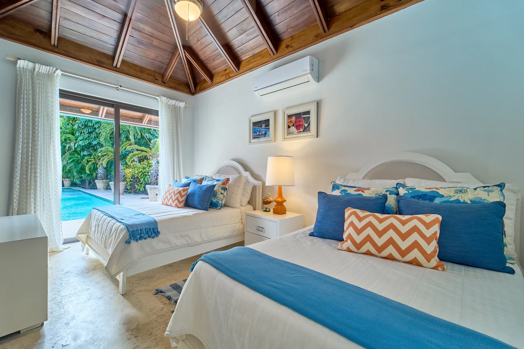 Un dormitorio luminoso con dos camas, ropa de cama azul y puertas de cristal hacia la vegetación.