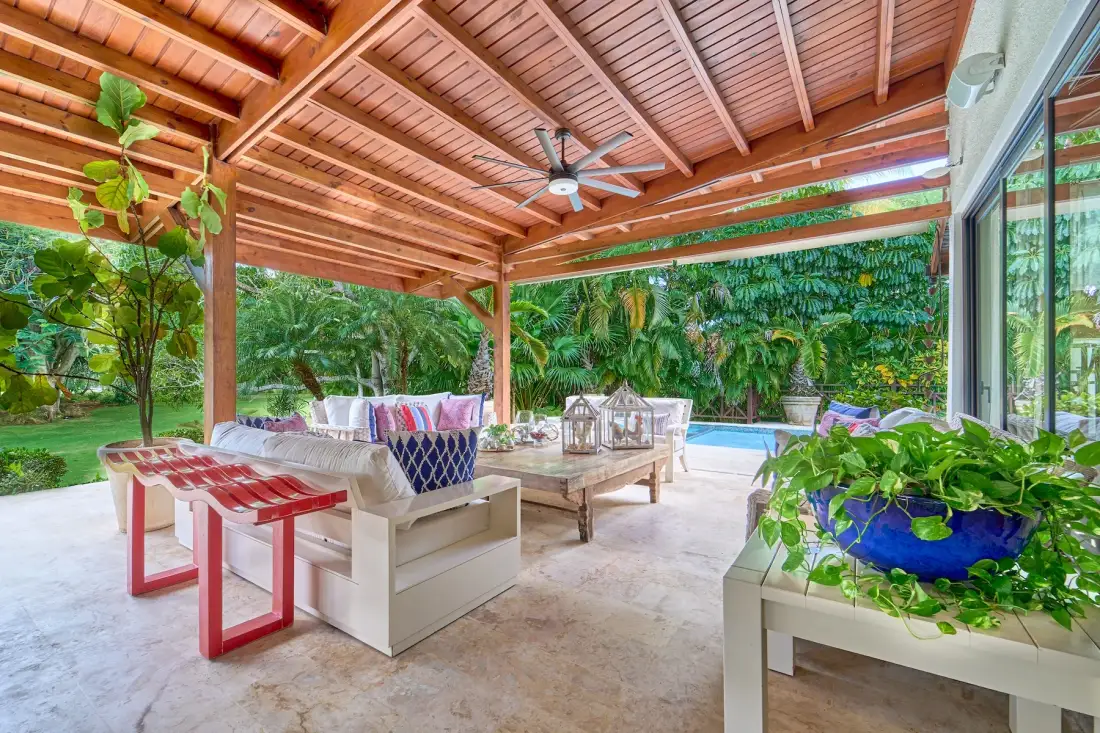 Una zona de estar exterior cubierta, con techo de madera y vegetación frondosa.
