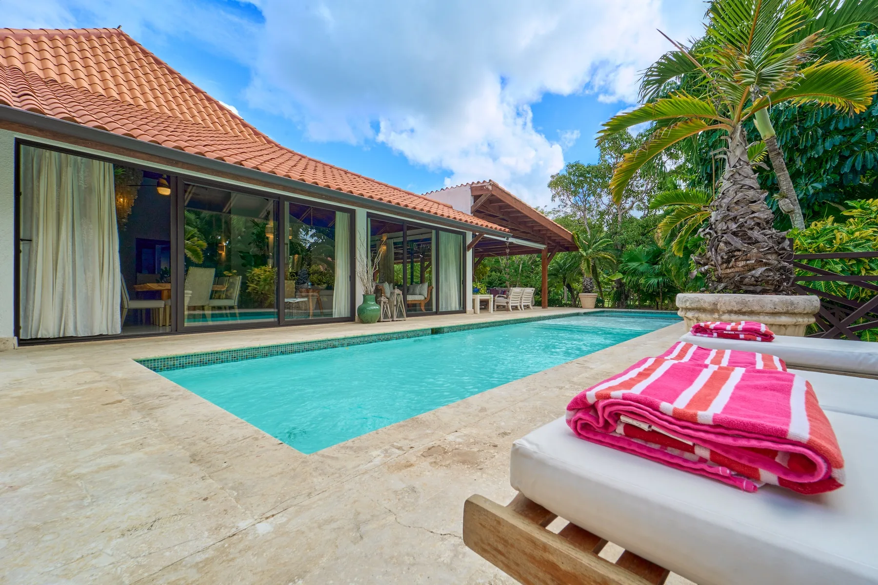 Una piscina junto a una casa con puertas de vidrio y plantas tropicales.