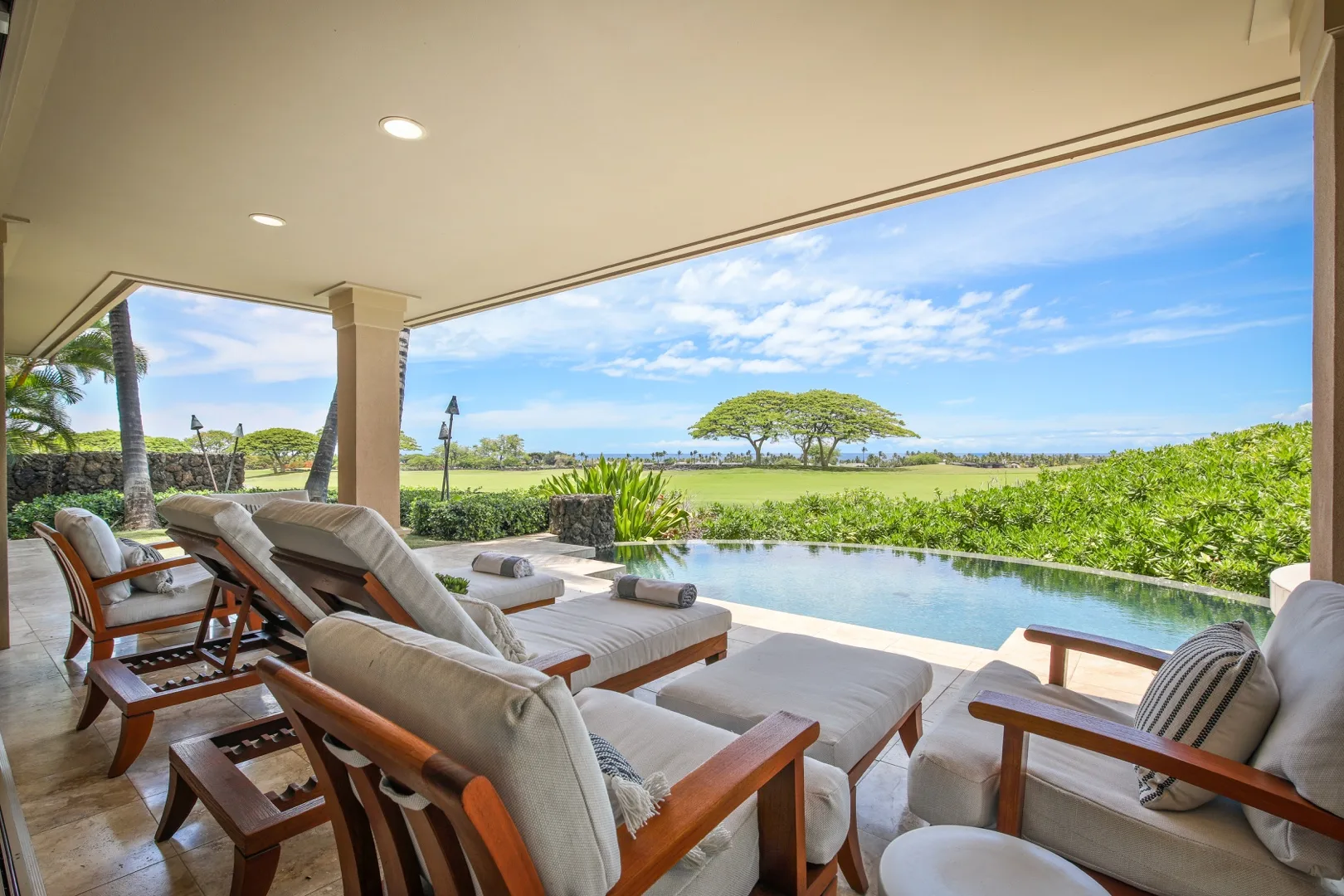 A covered patio with cushioned seating overlooks a pool and green landscape.