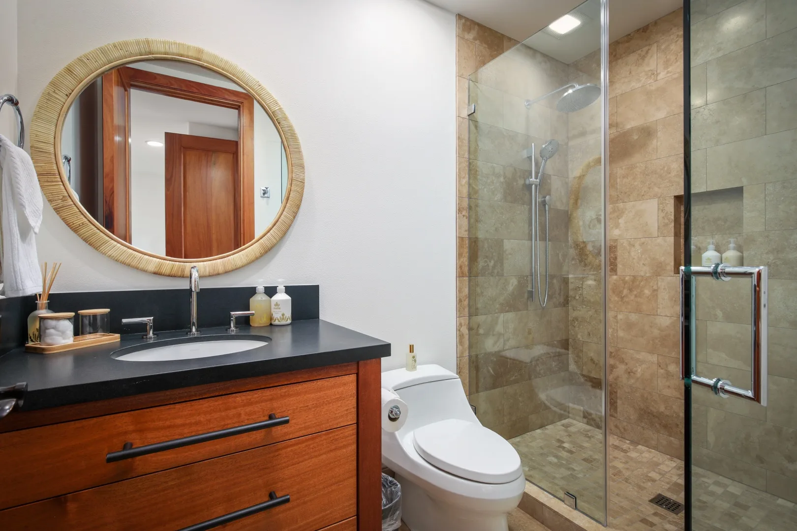 A bathroom with a vanity, oval mirror, toilet, and glass shower.