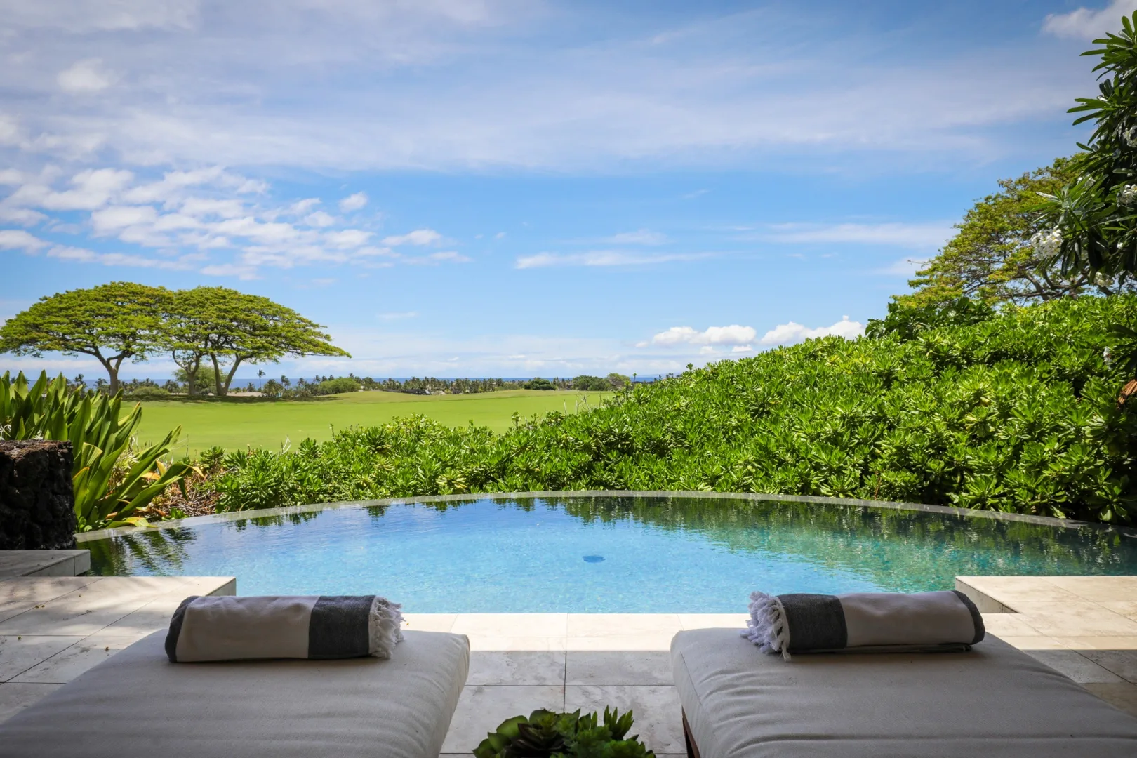 An outdoor swimming pool overlooks a green landscape beneath a partly cloudy sky.