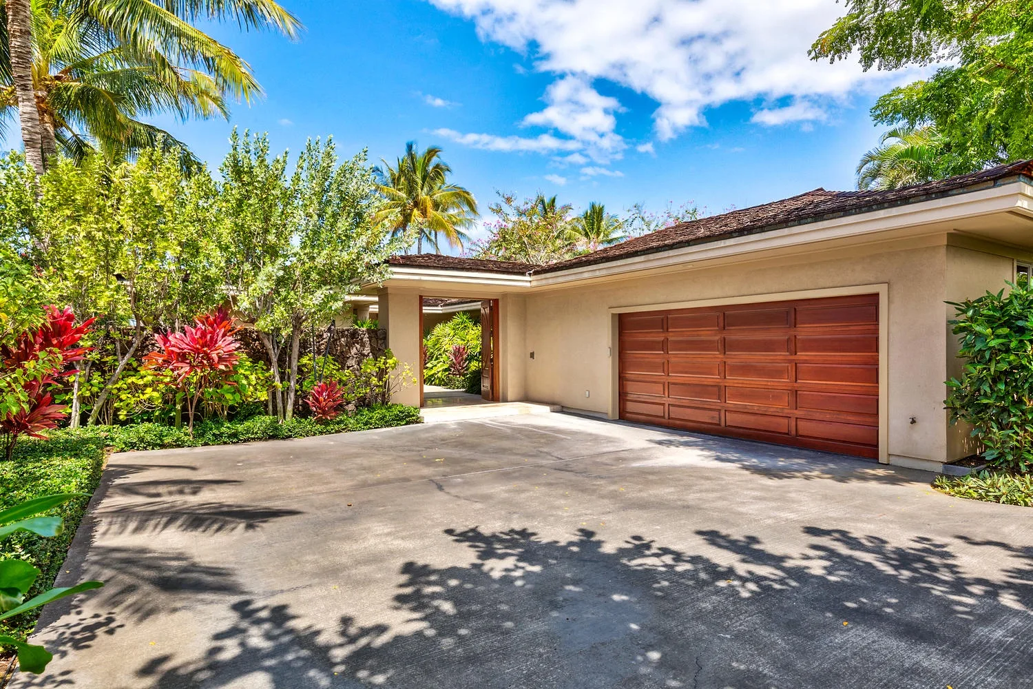 A house exterior with a broad driveway, wooden garage door, and tropical landscaping.
