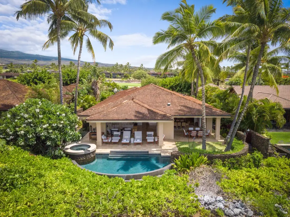 A tropical house with a tiled roof overlooks a swimming pool amid palm trees.