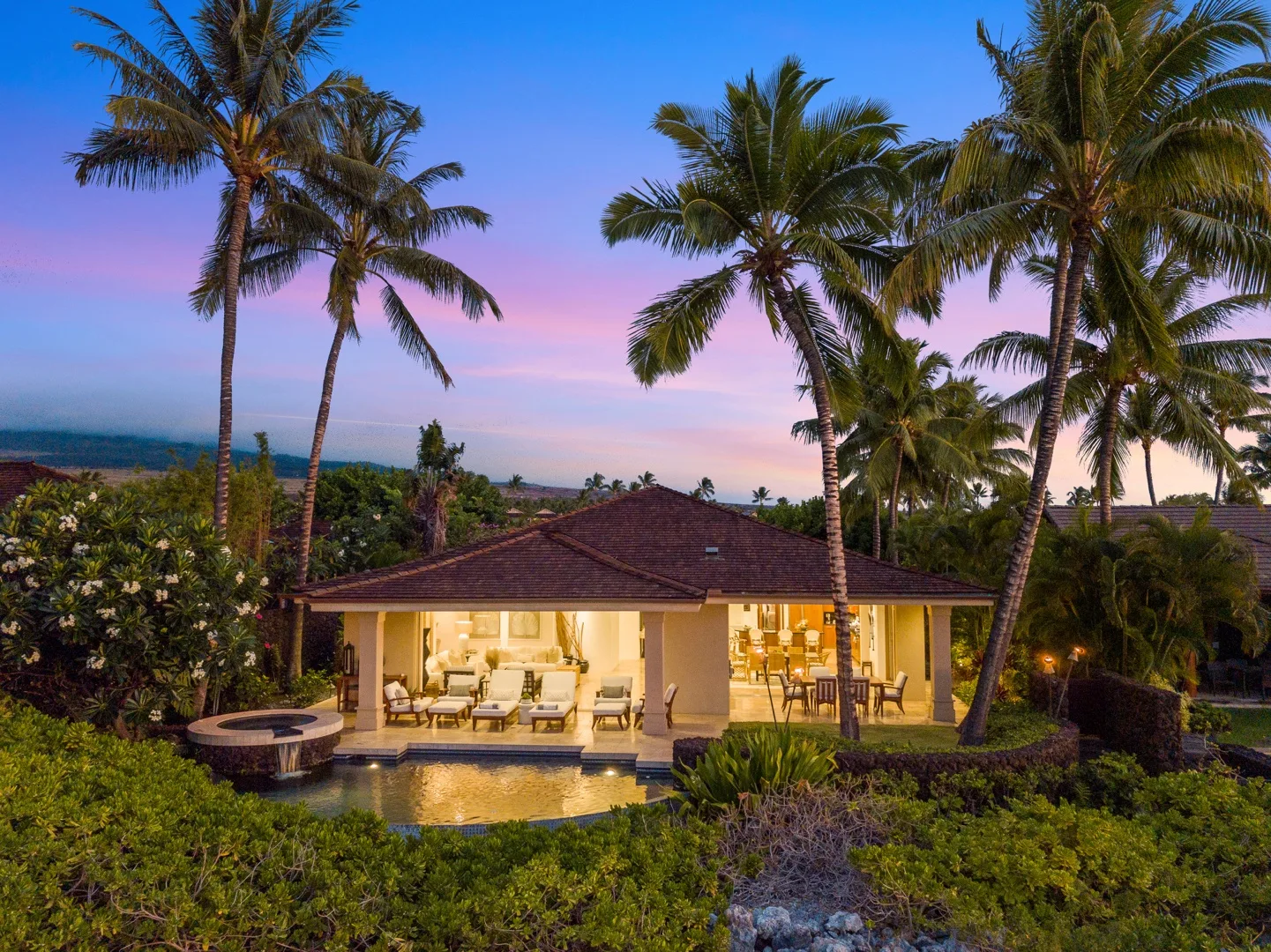 A tropical villa sits among palm trees beside a swimming pool at dusk.