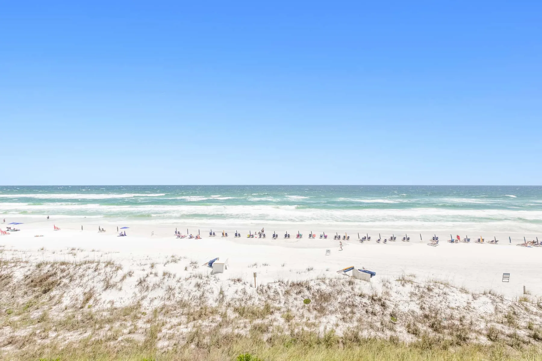 A sandy beach with dune grass, ocean waves, and people along the shore.