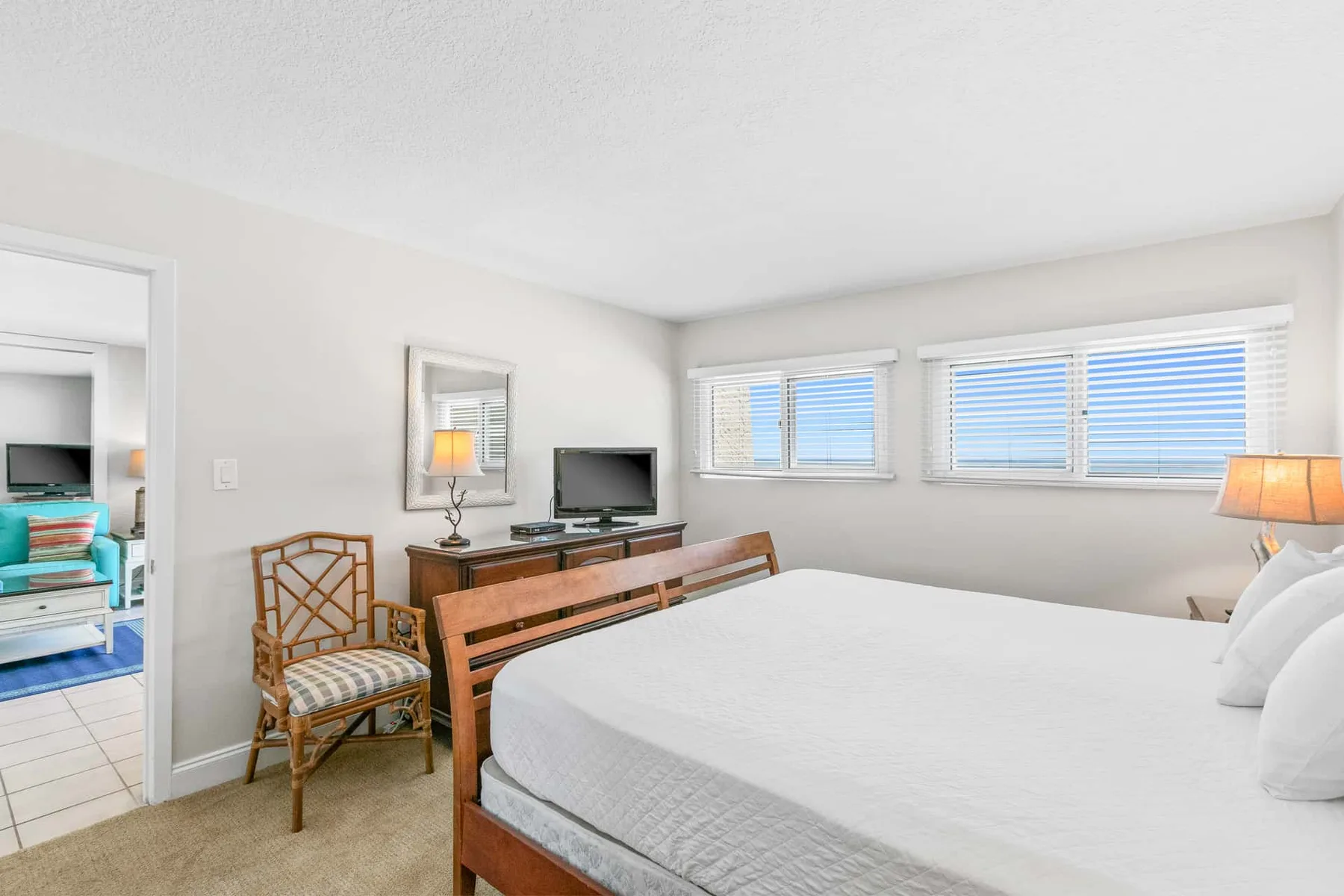 A bright bedroom with a neatly made bed, a desk, and windows with blinds.