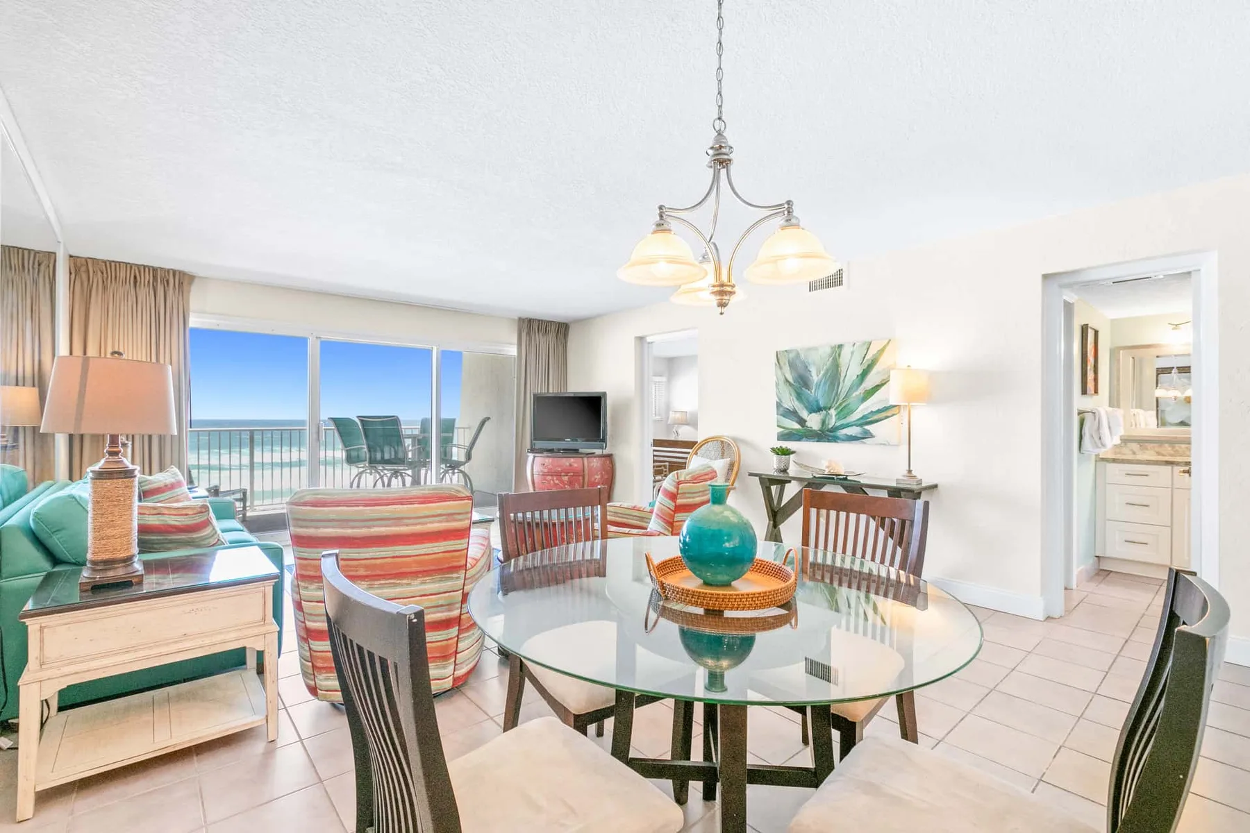 A bright dining and living area has a glass table, seating, and large windows overlooking the ocean.