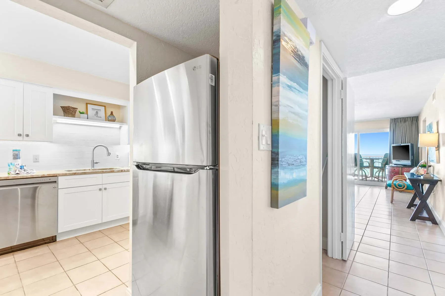 A kitchen with white cabinets and a refrigerator opens toward a bright living area.
