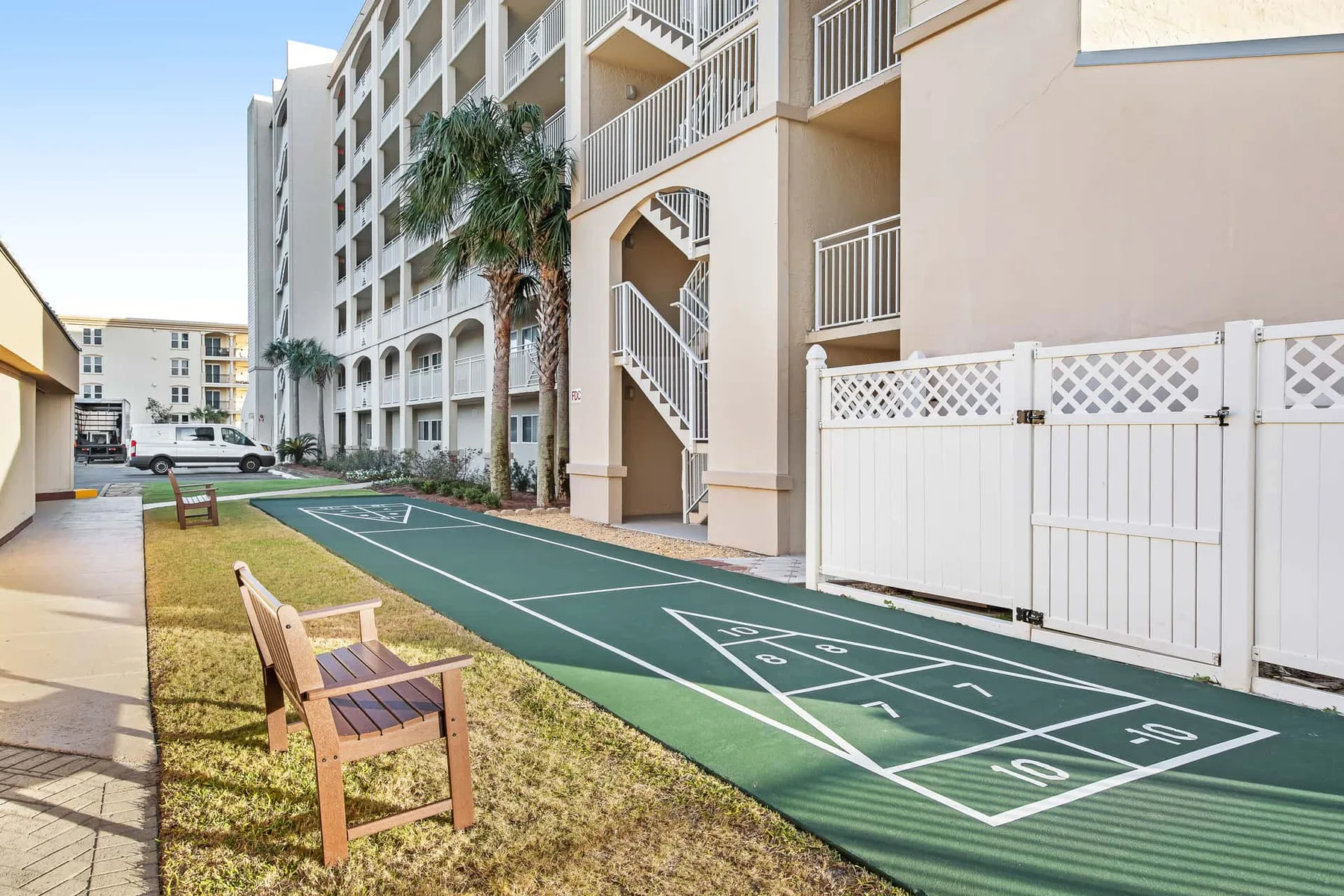 An outdoor shuffleboard court runs beside a multi-story building and a grassy seating area.