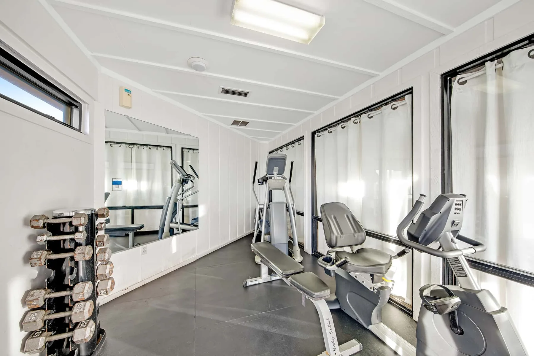 A bright exercise room with cardio machines and a rack of weights.