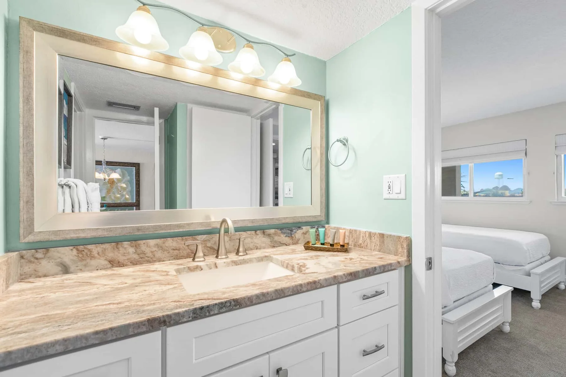 A bathroom vanity with a wide mirror opens onto a bedroom with white bedding.