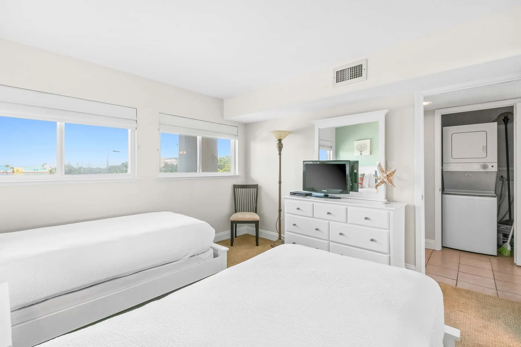 A bright bedroom with two beds, windows, and a dresser with a television.