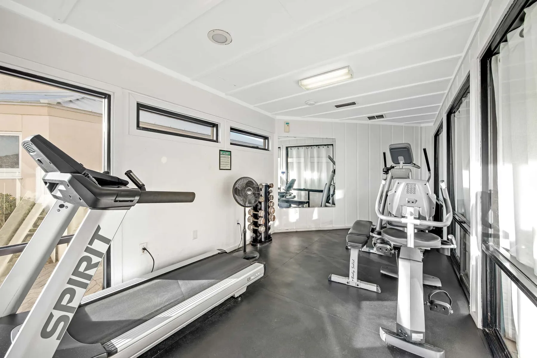 A bright gym room with treadmills, weight equipment, and large windows.