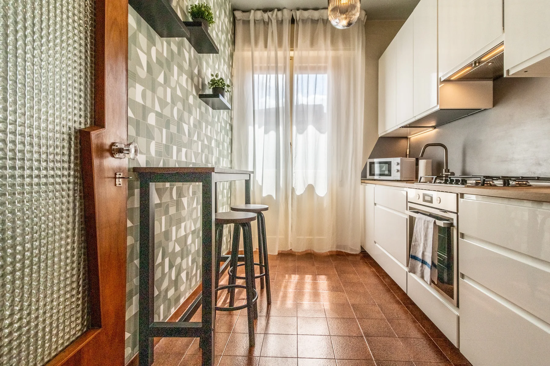 Una cocina estrecha con gabinetes blancos, una encimera y una ventana con cortinas transparentes.