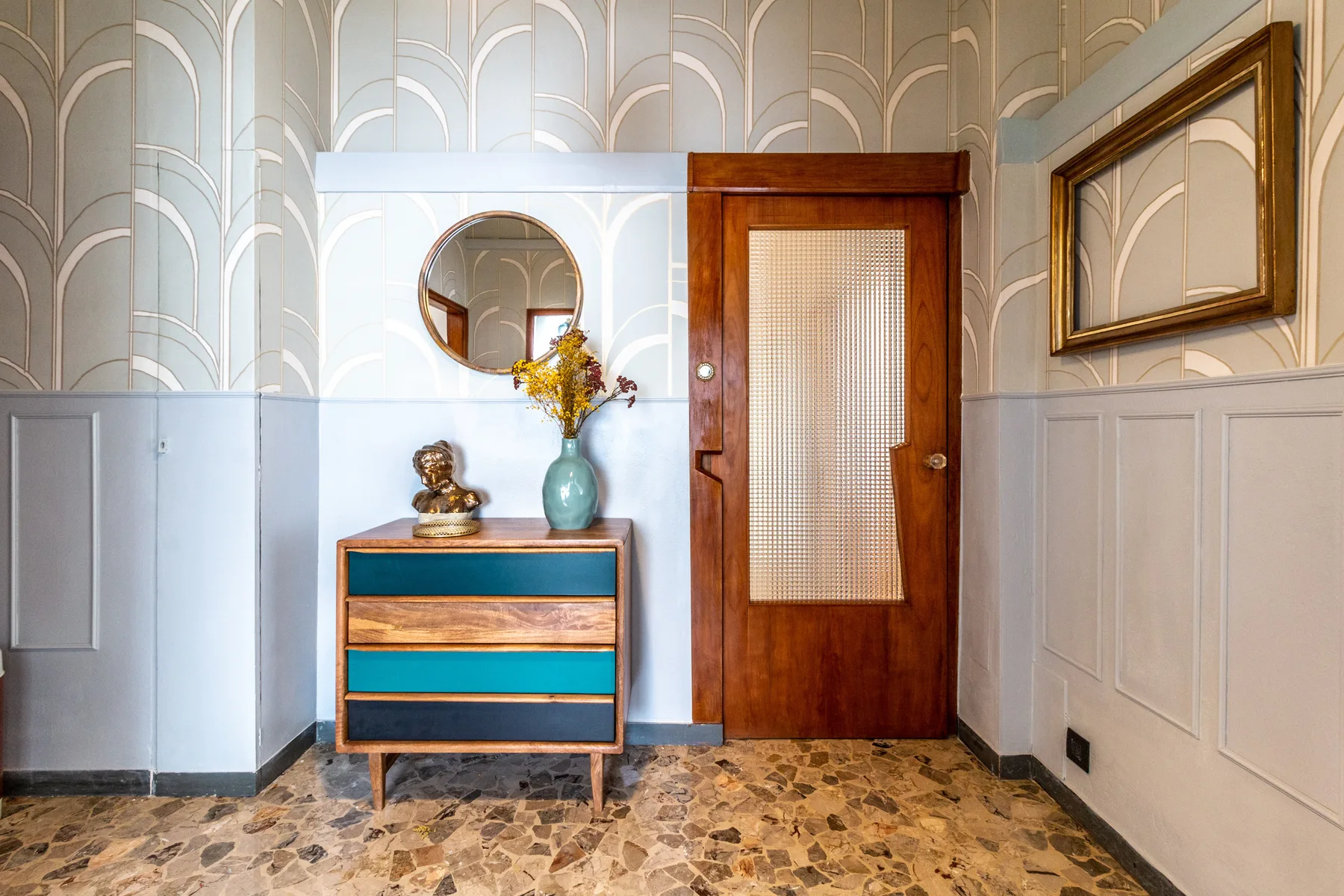 Un pasillo con una puerta de madera, una cómoda colorida y un espejo redondo.