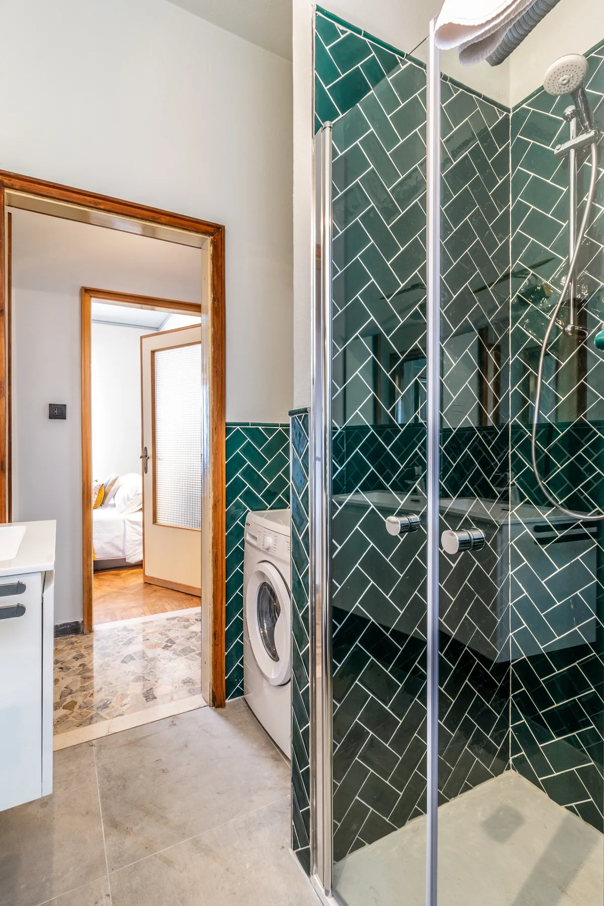 Un baño alicatado con ducha de vidrio, lavadora y una puerta hacia un dormitorio.