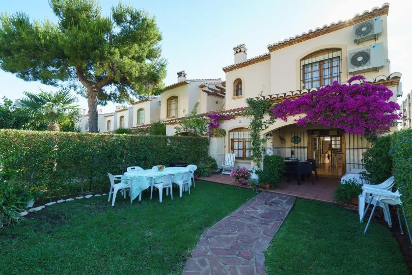 Un patio ajardinado junto a una casa de estilo mediterráneo, con asientos al aire libre y enredaderas con flores moradas.