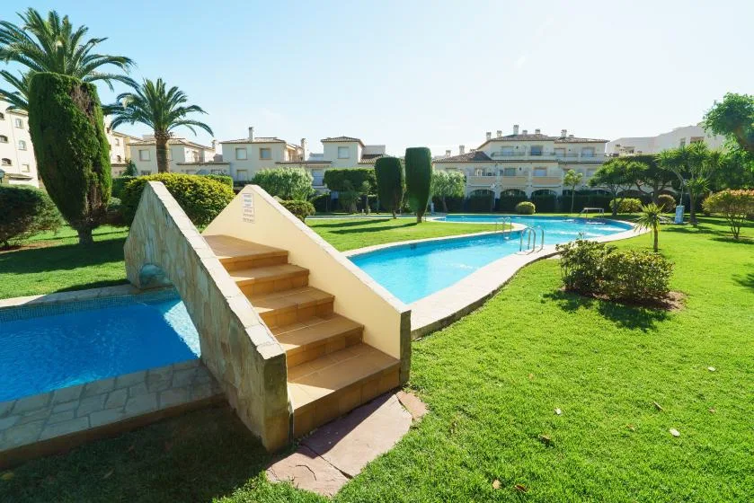 Una piscina con escalones está en un jardín rodeado de viviendas.