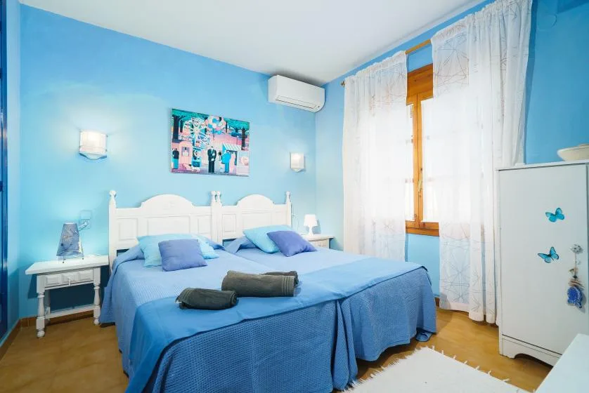 Un dormitorio luminoso con ropa de cama azul, muebles blancos y una ventana con cortinas.