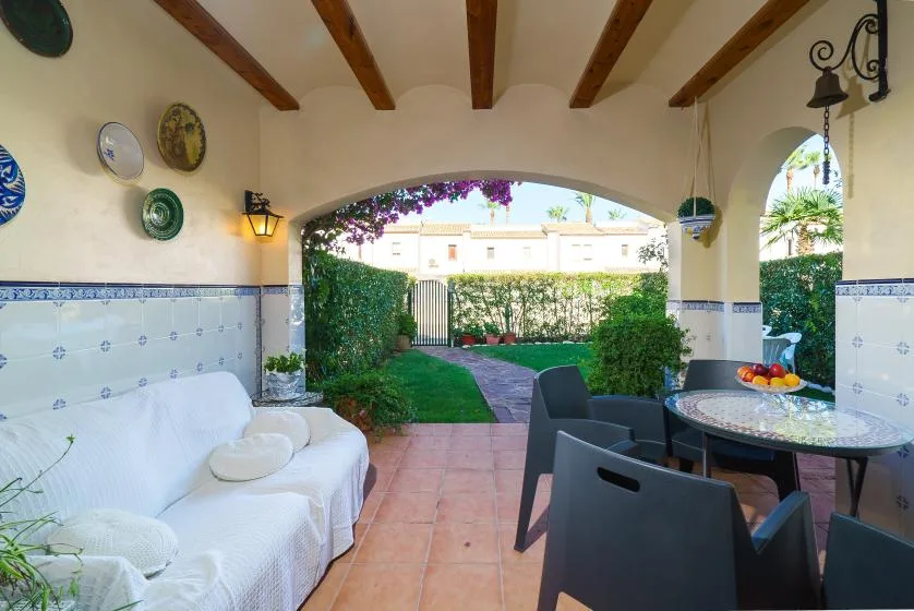 Un patio cubierto con asientos al aire libre, una mesa y un jardín al fondo.