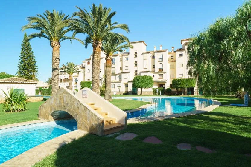Una zona ajardinada con piscina incluye un pequeño puente de piedra y palmeras.