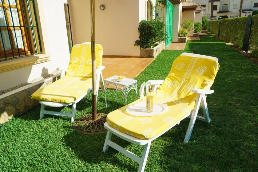 Un patio exterior con césped, asientos acolchados amarillos y una mesita junto al edificio.