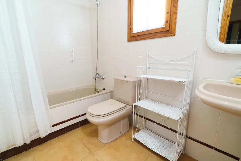 Un baño con bañera, inodoro y una estantería metálica blanca junto a una ventana.
